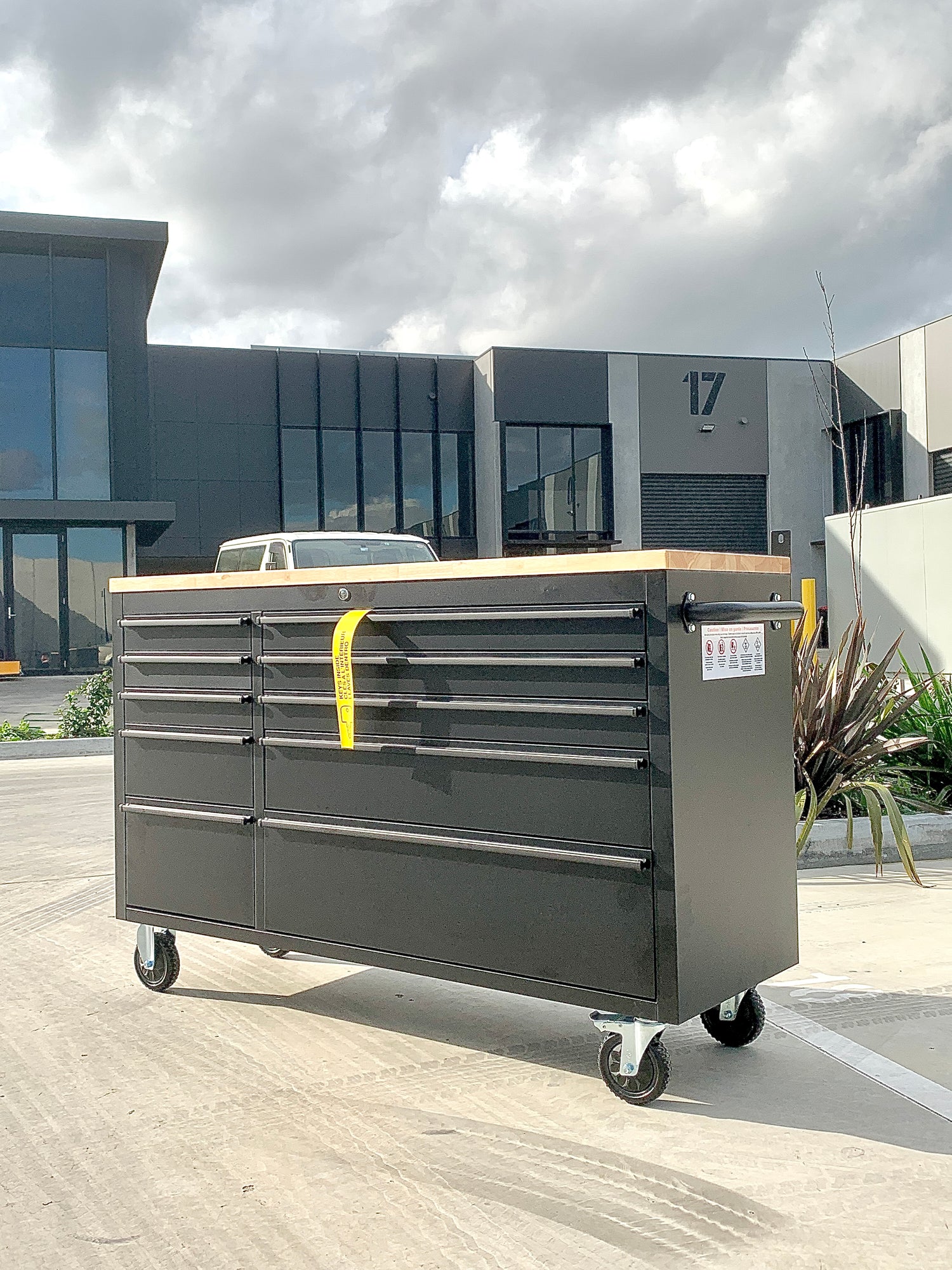 Black rolling tool cabinet with a wooden top and drawers, positioned in front of modern industrial buildings